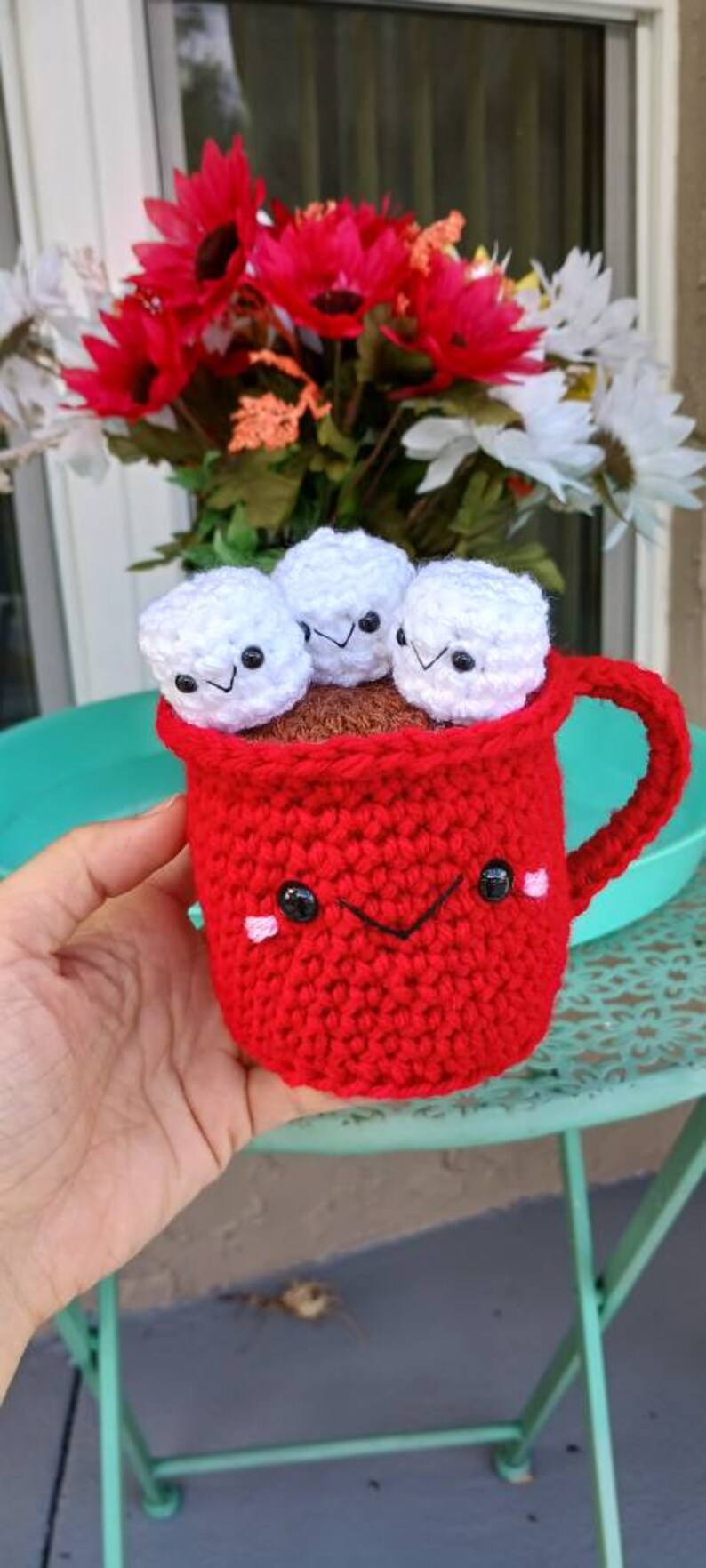 May include: A red crocheted mug with a smiling face and three white crocheted marshmallows inside. The mug has a handle and is sitting on a green table.
