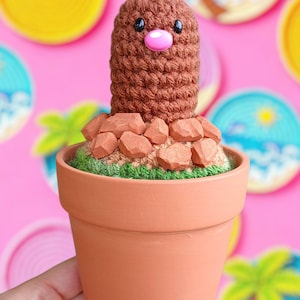 May include: A handmade crochet figure of a brown creature with a pink nose, resembling a Diglett, in a terracotta pot. The pot is filled with green yarn and brown rocks. The background features colorful, circular decorations.