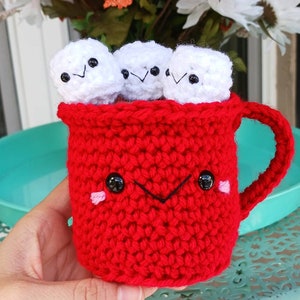 May include: A red crocheted mug with a smiling face and black eyes. The mug is filled with four white crocheted marshmallows with black eyes and smiling faces.