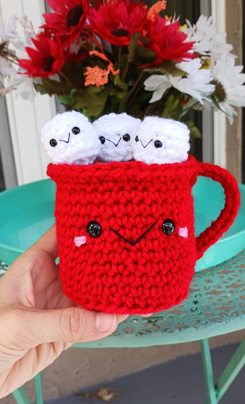 May include: A red crocheted mug with a smiling face and black eyes. The mug is filled with four white crocheted marshmallows with black eyes and smiling faces.