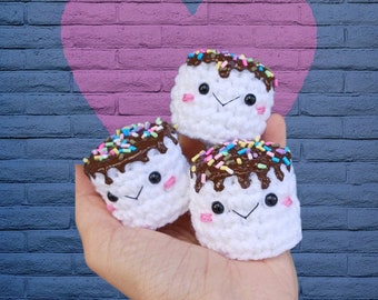 May include: Three crocheted marshmallow-shaped treats with chocolate frosting and colorful sprinkles. Each marshmallow has a cute face with eyes, a mouth, and rosy cheeks.