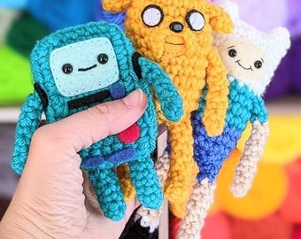 May include: Three crocheted plush toys, a blue robot, a yellow dog, and a white character with a blue hat, are held in a hand. The toys are all smiling and have large eyes.