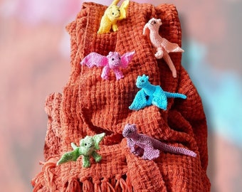 May include: A collection of crocheted dragon toys in various colors, including pink, blue, yellow, and green, are displayed on a textured orange blanket.