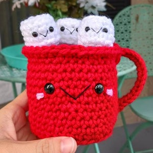 May include: A red crocheted mug with a smiling face and black eyes. The mug is filled with four white crocheted marshmallows.