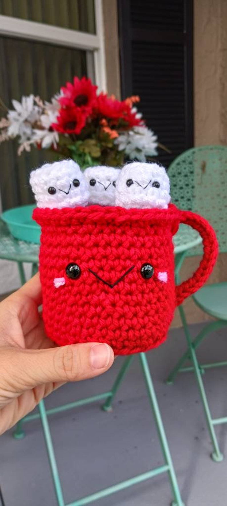 May include: A red crocheted mug with a smiling face and black eyes. The mug is filled with four white crocheted marshmallows.