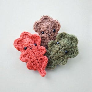 May include: Three crocheted leaf-shaped ornaments in shades of red, brown, and green. Each leaf has a smiling face with black bead eyes and a stitched mouth. The leaves are arranged on a white background.