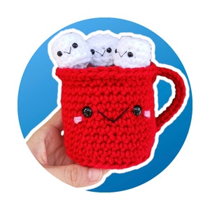 May include: A red crocheted mug with a white crocheted handle and three white crocheted marshmallows inside. The mug has a smiling face with black eyes and a pink blush.