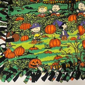 Puede incluir: Manta de forro polar verde con un diseño de Halloween con calabazas naranjas y personajes de dibujos animados. La manta tiene un borde de flecos anudados con tela negra y verde. Los personajes visten disfraces de Halloween.
