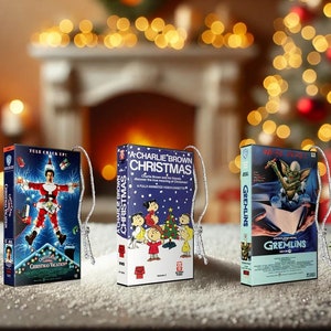 May include: Three Christmas VHS tape ornaments. One features the film "Christmas Vacation", another "A Charlie Brown Christmas", and the third "Gremlins". Each ornament has a silver string for hanging. The background is blurred with festive lights.