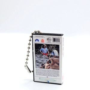 K&ouml;nnte beinhalten: Ein Miniatur-VHS-Kassetten-Schl&uuml;sselanh&auml;nger mit dem Film "Freitag der 13.". Der Anh&auml;nger hat eine silberne Kugelkette und einen Metallverschluss. Das Cover des Films ist zu sehen, zusammen mit Text auf der R&uuml;ckseite.