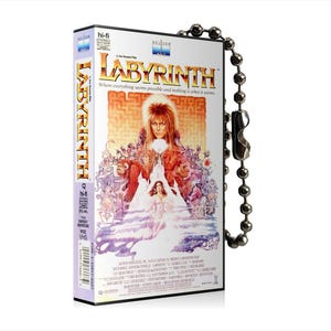 Puede incluir: Un llavero con forma de cinta VHS que representa la película Labyrinth. El llavero es plateado y tiene una imagen en blanco y negro de la portada de la película. La portada presenta el título de la película "Labyrinth" en letras grandes y en negrita y una imagen de David Bowie como Jareth el Rey Goblin.