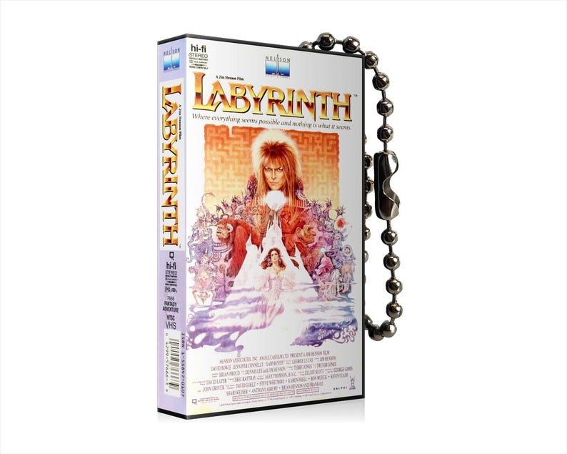 May include: A VHS tape keychain featuring the film Labyrinth. The keychain is silver and has a black and white image of the film's cover art. The cover art features the film title "Labyrinth" in large, bold letters and a picture of David Bowie as Jareth the Goblin King.