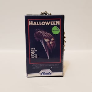 K&ouml;nnte beinhalten: Ein Miniatur-VHS-Kassetten-Schl&uuml;sselanh&auml;nger mit dem Filmplakat von "Halloween". Das Poster zeigt ein Messer und einen K&uuml;rbis vor einem dunkelblauen Hintergrund. Der Text "The Night He Came Home!" ist ebenfalls sichtbar.