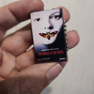 K&ouml;nnte beinhalten: Kleiner rechteckiger Schl&uuml;sselanh&auml;nger mit dem Filmplakat f&uuml;r "Das Schweigen der L&auml;mmer". Das Plakat zeigt das Gesicht einer Frau mit einer Motte &uuml;ber ihrem Mund. Der Text "the silence of the lambs" ist rot.