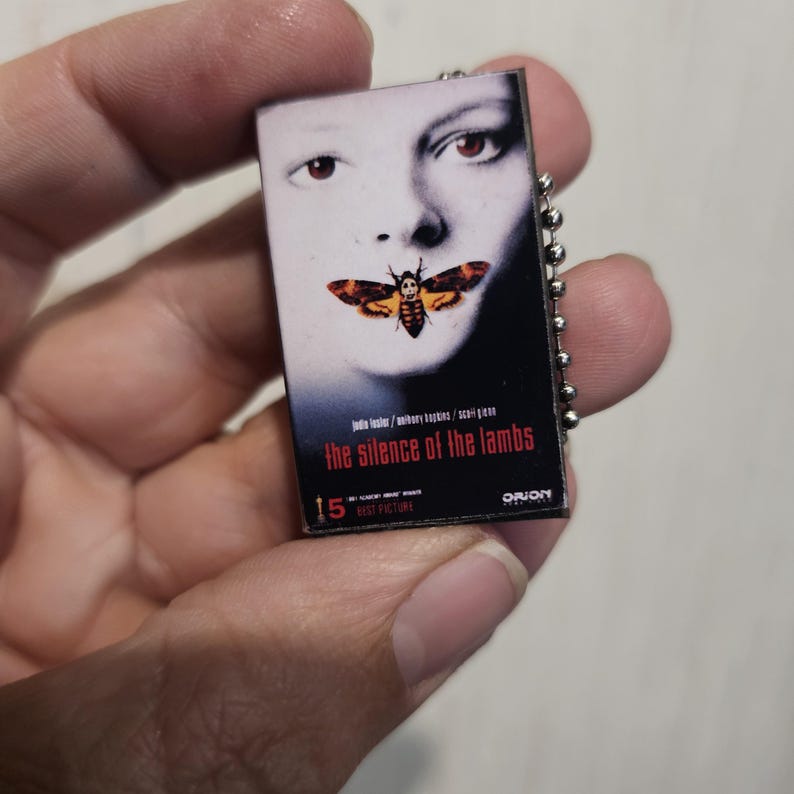 K&ouml;nnte beinhalten: Kleiner rechteckiger Schl&uuml;sselanh&auml;nger mit dem Filmplakat f&uuml;r "Das Schweigen der L&auml;mmer". Das Plakat zeigt das Gesicht einer Frau mit einer Motte &uuml;ber ihrem Mund. Der Text "the silence of the lambs" ist rot.