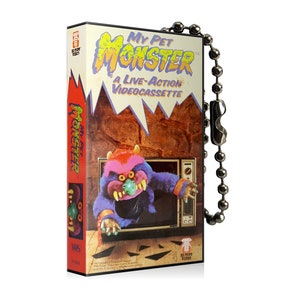 May include: A novelty VHS tape-shaped keyring with the text "My Pet Monster: A Live-Action Videocassette." The design includes a cartoon monster emerging from a television set. The keyring has a silver ball chain.