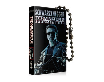 Könnte beinhalten: Ein schwarzer und silberner Schlüsselanhänger mit einem Terminator 2: Judgment Day VHS-Kassetten-Design mit dem Bild von Arnold Schwarzenegger auf der Vorderseite. Der Schlüsselanhänger hat eine silberne Kette.