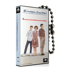 May include: A VHS tape keychain featuring the movie "Sixteen Candles". The keychain is silver with a black and white image of the movie's main characters on the front. The text "Sixteen Candles" is printed in blue on the front of the keychain.