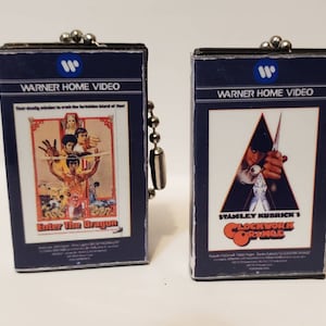 May include: Two miniature VHS cassette keychains, one featuring the movie "Enter the Dragon" and the other featuring "A Clockwork Orange". Both keychains are blue and feature the Warner Home Video logo.