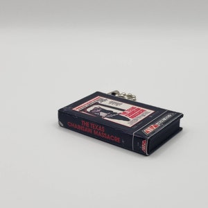 K&ouml;nnte beinhalten: Eine kleine, rechteckige, dunkelblaue VHS-Kassetten-Replik mit dem Titel "THE TEXAS CHAINSAW MASSACRE" in wei&szlig;er und roter Schrift. Das Band hat eine silberne Kette. Das Bild zeigt einen Mann mit einer Kettens&auml;ge.