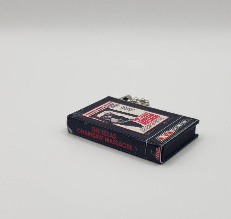 K&ouml;nnte beinhalten: Eine kleine, rechteckige, dunkelblaue VHS-Kassetten-Replik mit dem Titel "THE TEXAS CHAINSAW MASSACRE" in wei&szlig;er und roter Schrift. Das Band hat eine silberne Kette. Das Bild zeigt einen Mann mit einer Kettens&auml;ge.