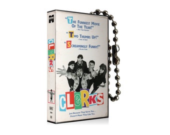 Könnte beinhalten: Ein Schlüsselanhänger in Form einer VHS-Kassette mit dem Film "Clerks" und einer Gruppe von Personen auf dem Cover. Der Text auf der Kassette lautet "The Funniest Movie of the Year!" "Two Thumbs Up!" "Screamingly Funny!" und "Clerks".