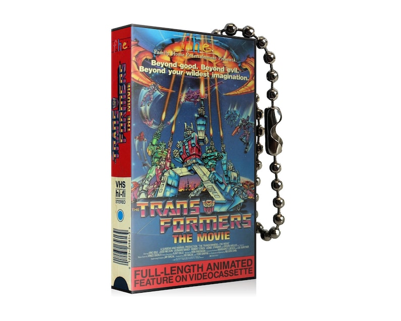 May include: A vintage VHS cassette tape keychain featuring the Transformers: The Movie artwork. The keychain is red, blue, and yellow with a silver chain. The text on the cassette reads "Transformers: The Movie" and "Full-length animated feature on videocassette".