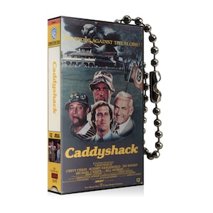 May include: A VHS tape case with a black background and a yellow title card that reads "Caddyshack". The case features a photo of four men from the movie, including Chevy Chase, Rodney Dangerfield, Ted Knight, and Bill Murray. The text "The Snobs Against The Slobs!" is printed in white on the top of the case.