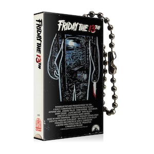 Puede incluir: Un llavero de cinta VHS negra con una cadena plateada. La parte delantera de la cinta presenta el cartel de la película "Viernes 13" con una silueta de Jason Voorhees sosteniendo un machete.