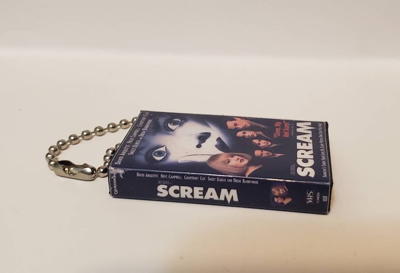 K&ouml;nnte beinhalten: Ein Miniatur-VHS-Kassetten-Schl&uuml;sselanh&auml;nger mit dem Film "Scream". Das Cover zeigt eine Nahaufnahme eines Gesichts mit weit aufgerissenen Augen und anderen Darstellern. Der Schl&uuml;sselanh&auml;nger hat eine silberne Kugelkette und einen Verschluss.