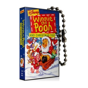 May include: A rectangular Winnie the Pooh and Christmas Too VHS tape with a silver ball chain. The cover features Winnie the Pooh dressed as Santa Claus on a sled with other characters in a snowy scene. The title is in red and yellow.