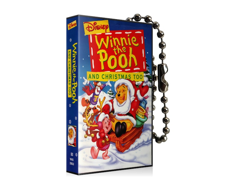 May include: A rectangular Winnie the Pooh and Christmas Too VHS tape with a silver ball chain. The cover features Winnie the Pooh dressed as Santa Claus on a sled with other characters in a snowy scene. The title is in red and yellow.