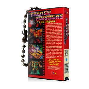 May include: A red and black VHS tape keychain with a silver chain. The front of the tape features a cartoon image of the Transformers logo and characters. The back of the tape has text describing the movie, including the title "Transformers: The Movie" and the run time of 86 minutes.