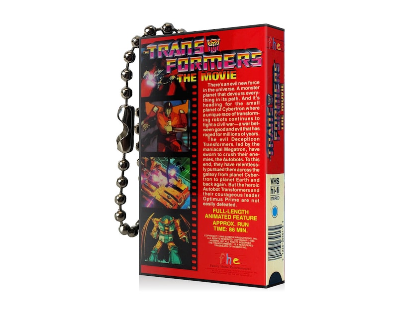 May include: A red and black VHS tape keychain with a silver chain. The front of the tape features a cartoon image of the Transformers logo and characters. The back of the tape has text describing the movie, including the title "Transformers: The Movie" and the run time of 86 minutes.
