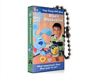 Puede incluir: Una caja de DVD azul y blanca con una imagen de Blue de Blue's Clues en la parte delantera. La caja tiene el texto "Blue's Discoveries" y "Play Along With Blue". La caja está unida a una cadena plateada.