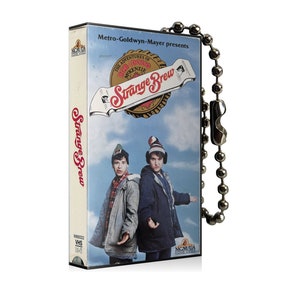 May include: A VHS tape of the movie "Strange Brew" featuring a blue sky background and two people wearing winter hats and coats. The tape has a silver ball chain attached. The movie title is in red and white.