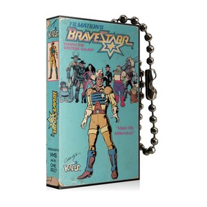 May include: A vintage VHS tape featuring the animated series "BraveStarr: Taming the Western Galaxy." The tape has a light blue cover with cartoon illustrations of characters. A silver ball chain is attached to the tape.