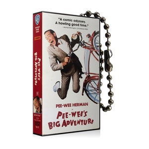 May include: A VHS tape keychain featuring Pee-wee Herman's Big Adventure. The keychain is red and white with a picture of Pee-wee Herman on a bicycle. The text "A comic odyssey. A howling good time." is printed on the keychain.