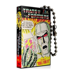 May include: A Transformers VHS tape keychain. The box art features a comic book style illustration of a robot character with the text "The Transformers" and "Volume 6". The keychain has a silver ball chain.