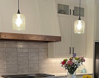 May include: A kitchen island with a dark gray countertop and three black stools, featuring a wooden base with open shelving. A white tray holds a flower vase and a small bowl of lemons. Three glass pendant lights hang above the island.