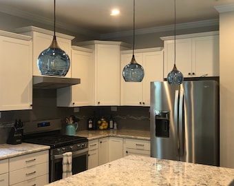 May include: Three pendant lights with rounded, translucent blue glass shades and dark metal hardware suspended from the ceiling above a kitchen island, featuring white cabinetry, a stainless steel refrigerator, and a granite countertop.