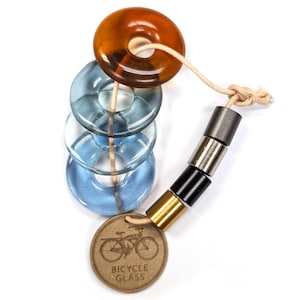 May include: A unique bicycle-themed accessory featuring a series of transparent, circular glass beads in shades of amber and blue. The beads are strung together with a natural cord, and a small, round tag with a bicycle illustration and the text "BICYCLE GLASS" is attached.