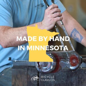 May include: A person is working with a glassblowing torch to create a glass bottle. The bottle is red and clear. The person is wearing a blue shirt and has a tattoo on their arm. The text "MADE BY HAND IN MINNESOTA" is displayed on a yellow outline of the state of Minnesota. The text "BICYCLE GLASS CO." is displayed below a bicycle icon.