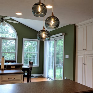 May include: Three glass globe pendant lights hang from a ceiling fixture in a kitchen. The globes are a dark teal color with a rippled texture. The lights are hanging over a kitchen island with a brown countertop.