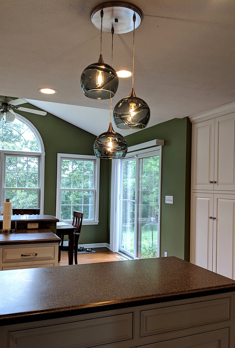 May include: Three glass globe pendant lights hang from a ceiling fixture in a kitchen. The globes are a dark teal color with a rippled texture. The lights are hanging over a kitchen island with a brown countertop.