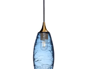 May include: A pendant light fixture featuring a cylindrical blue glass shade with a textured, rippled surface pattern. The shade is suspended by a black cord and attached to a metallic gold-toned cap that houses a single clear light bulb.