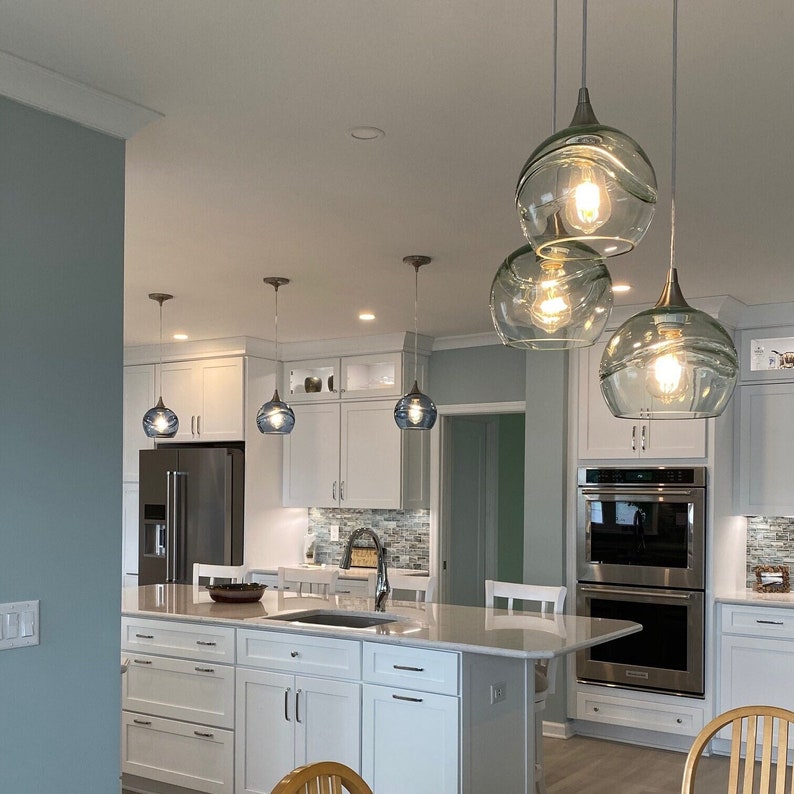 May include: A modern kitchen with white cabinets and a large island. The island has a white countertop and a stainless steel sink. There are three blue glass pendant lights hanging over the island. The kitchen has a light blue wall and a stainless steel double oven.