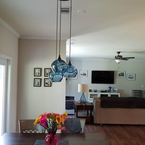May include: A three-light pendant light fixture with blue glass shades hangs from the ceiling in a dining room. The shades are round and have a textured surface. A vase of flowers sits on a wooden table in the foreground.