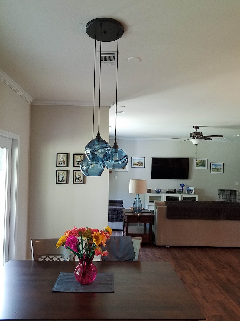 May include: A three-light pendant light fixture with blue glass shades hangs from the ceiling in a dining room. The shades are round and have a textured surface. A vase of flowers sits on a wooden table in the foreground.