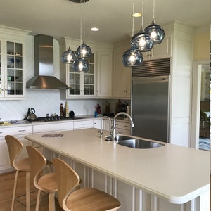 May include: A white kitchen island with a white countertop and a stainless steel sink. Three wooden bar stools with chrome legs are positioned around the island. A set of five blue glass pendant lights hang above the island.